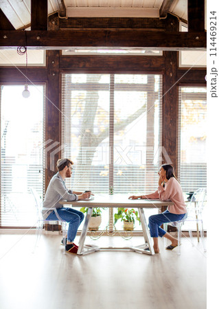 Couple sharing ideas, sitting opposite near big window. Couple sharing ideas, sitting opposite near big window. 114469214