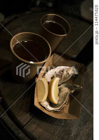 Opened oyster with lemon, pear cider in paper cups on wooden table barrel in dim light. Seafood dining, rustic ambiance, gourmet cuisine, beverage pairing, elegant setting, culinary scene Opened oyster with lemon, pear cider in paper cups on wooden table barrel in dim light. Seafood dining, rustic ambiance, gourmet cuisine, beverage pairing, elegant setting, culinary scene 114475820