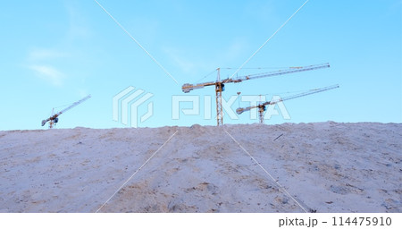 Three construction cranes tower over a mound of sand against a backdrop of blue sky. Perfect for illustrating construction progress and industrial development Three construction cranes tower over a mound of sand against a backdrop of blue sky. Perfect for illustrating construction progress and industrial development 114475910