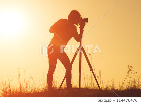 Silhouette of woman photographer shooting landscape with camera on tripod at sunset time in the field Silhouette of woman photographer shooting landscape with camera on tripod at sunset time in the field 114476476