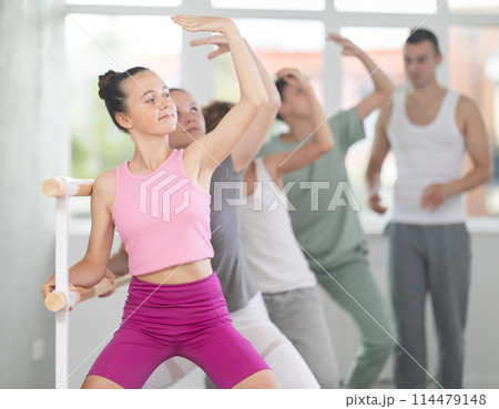 Teenagers doing ballet at group training session in the studio performs demi plie near the ballet barre, standing in ballet stance Teenagers doing ballet at group training session in the studio performs demi plie near the ballet barre, standing in ballet stance 114479148