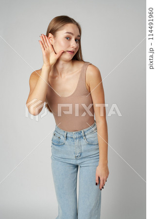 Young woman puts a hand to the ear to hear better on gray background Young woman puts a hand to the ear to hear better on gray background 114491960