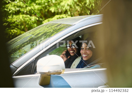 Two Muslim women wearing hijab converse on a smartphone while traveling together in a car through the  114492382
