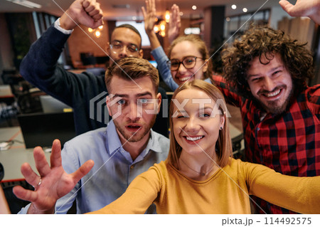 A diverse group of business professionals take a break from their tasks in a modern startup office to capture a creative selfie, showcasing teamwork and a vibrant workplace culture 114492575