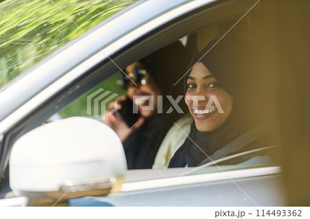 Two Muslim women wearing hijab converse on a smartphone while traveling together in a car through the  114493362