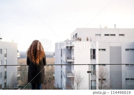 Rear view of woman with curly hair standing on terrace, enjoying cold autumn morning, sunny day. Speaking positive affirmations. 114498819