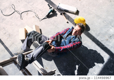 Male worker lying on the floor after fall from ladder, holding his knee. Work injury, accident in workplace. 114498967