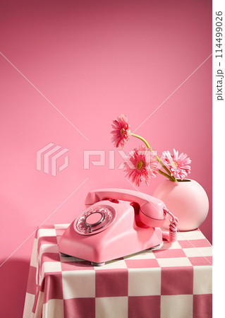 Pink background against a desk phone with pink color and a flower vase of pink gerbera daisy placed on a table has checkered pattern. Frontal shot with space for text 114499026