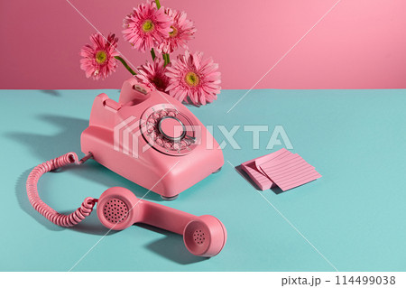 A desk phone with receiver placed on a flat blue surface, a few branches of pink gerbera flowers decorated on the back and a stack of sticky notes of the same color placed next to it. Copy space 114499038