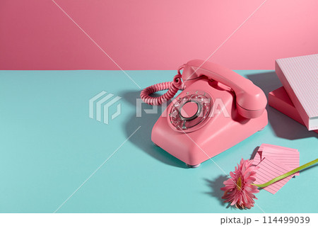Pink color desk phone, some books, sticky notes and a gerbera daisy branch displayed on the right side of blue table over pink background. Blank space on the left for showing product or adding text 114499039