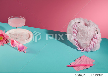 Some daily life accessories placed on blue table such as a light pink hand bag, sticky notes, a glasses, gerbera daisy and cocktail glass with pink liquid. Vacant space in center, high angle shot 114499040