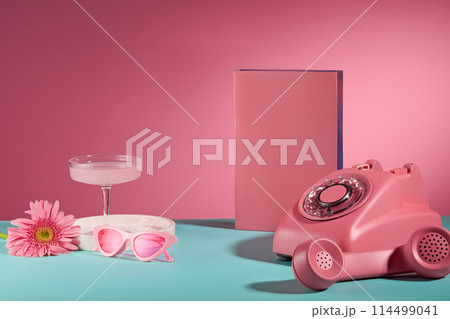 Life style pink item on blue table over pink background, a desk phone, pink pedestal, a glasses, cocktail glass and daisy gerbera. Space for adding text, front view 114499041