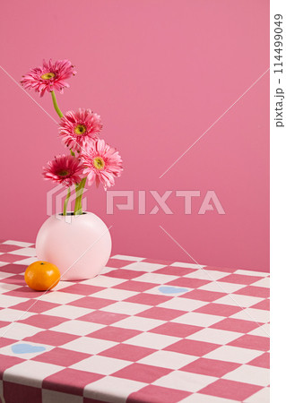 Advertising photo in pink color with a white flower vase of pink gerbera daisy next to an orange placed on the table covered with a checkered patterned towel, over pink background. Space for design 114499049