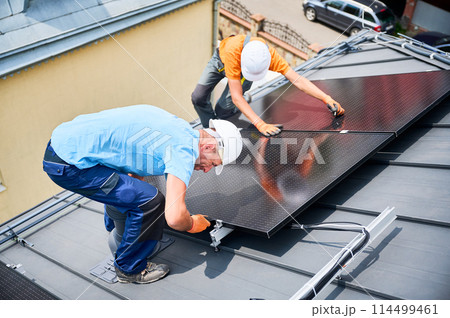 Workers building photovoltaic solar panel system on rooftop of house. Men technicians in helmets and gloves installing solar module with help of hex key outdoors. Renewable energy generation concept. 114499461