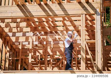 Carpenter constructing wooden frame, two-story house. Bearded man hammering nails with hammer, dressed in protective helmet and work coveralls. Concept of environmentally-friendly modern construction. Carpenter constructing wooden frame, two-story house. Bearded man hammering nails with hammer, dressed in protective helmet and work coveralls. Concept of environmentally-friendly modern construction. 114499506
