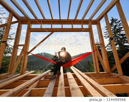 Man on construction site of wooden-framed house sits in hammock against backdrop of mountains and sunrise, sipping coffee. The concept pertains to modern ecological construction. 114499566