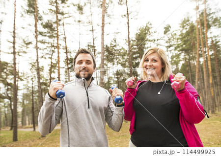 An overweight woman exercising outdoors with friend, fitness coach. Holding dumbbells. 114499925