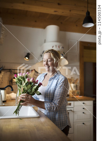 Beautiful mature woman arranging bouquet of roses in a vase, a hobby and relaxation. Older woman living alone, enjoying peaceful weekend day 114500334