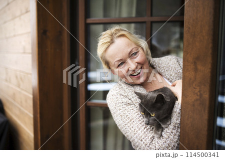 Beautiful mature woman at home, holding, petting her cat, looking through the window. Older woman living alone, enjoying peaceful weekend day 114500341
