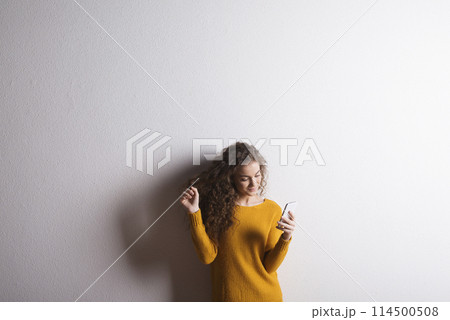 Portrait of a gorgeous teenage girl with curly hair, holding smartphone, scrolling. Studio shot, white background with copy space Portrait of a gorgeous teenage girl with curly hair, holding smartphone, scrolling. Studio shot, white background with copy space 114500508