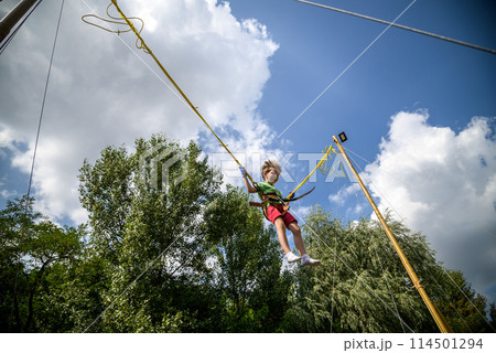 The boy is jumping on a bungee trampoline. A child with insurance and stretchable rubber bands hangs against the sky. The concept of happy childhood and games in the amusement park The boy is jumping on a bungee trampoline. A child with insurance and stretchable rubber bands hangs against the sky. The concept of happy childhood and games in the amusement park 114501294