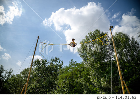 The boy is jumping on a bungee trampoline. A child with insurance and stretchable rubber bands hangs against the sky. The concept of happy childhood and games in the amusement park The boy is jumping on a bungee trampoline. A child with insurance and stretchable rubber bands hangs against the sky. The concept of happy childhood and games in the amusement park 114501362