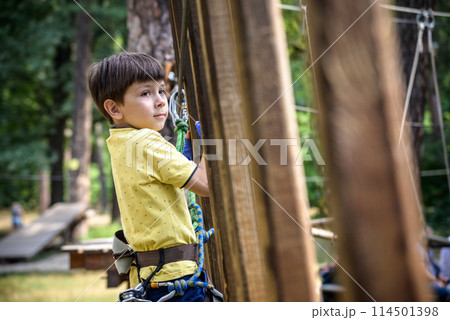 Strong excited young boy playing outdoors in rope park. Caucasian child dressed in casual clothes and sneakers at warm sunny day. Active leisure time with children concept Strong excited young boy playing outdoors in rope park. Caucasian child dressed in casual clothes and sneakers at warm sunny day. Active leisure time with children concept 114501398