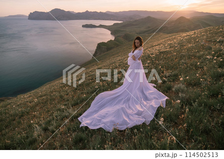 A woman in a long white dress stands on a hill overlooking a body of water 114502513