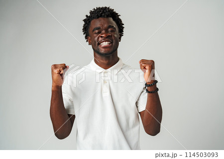 Young African Man in White Shirt Celebrates With Joyful Fist Pump on Grey Background Young African Man in White Shirt Celebrates With Joyful Fist Pump on Grey Background 114503093