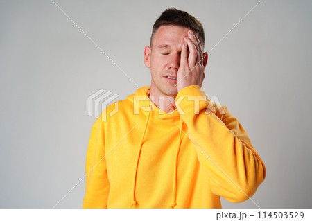 Portrait of young man in yellow hoodie with face palm gesture on gray background 114503529