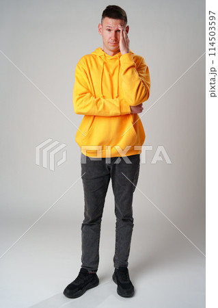 Portrait of young man in yellow hoodie with face palm gesture on gray background 114503597