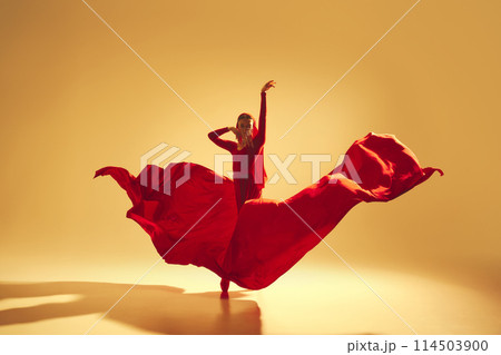 Artistic and deep performance Woman, female dancer in flowing red dress dancing against golden color background 114503900
