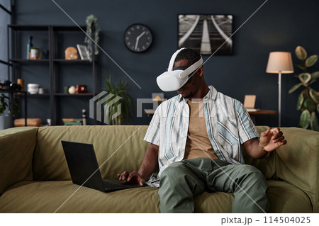 Young African American man sitting on sofa in living room setting VR headset using app on laptop, copy space Young African American man sitting on sofa in living room setting VR headset using app on laptop, copy space 114504025