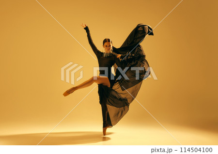 Artistic and expressive performance. Young woman, ballet dancer in black flowing attire dancing against golden background Artistic and expressive performance. Young woman, ballet dancer in black flowing attire dancing against golden background 114504100