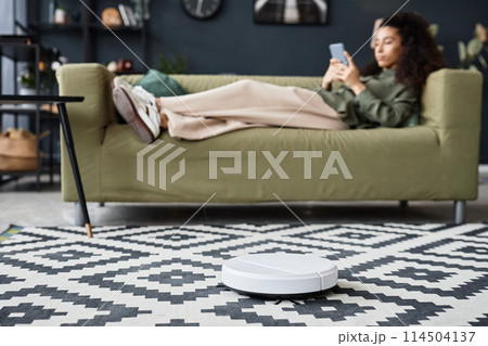 Selective focus shot of robotic vacuum cleaner cleaning floor in living room while young woman relaxing on couch 114504137