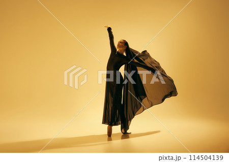 Desert freedom. Elegant and artistic young woman in black attire with veil dancing, performing ballet against sand color background Desert freedom. Elegant and artistic young woman in black attire with veil dancing, performing ballet against sand color background 114504139