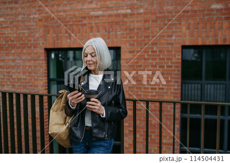Portrait of stylish mature woman with gray hair on city street. Older woman in leather jacket with soft smile. 114504431