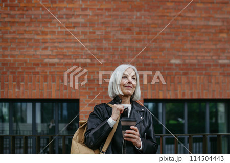 Portrait of stylish mature woman with gray hair on city street. Older woman in leather jacket with soft smile. Portrait of stylish mature woman with gray hair on city street. Older woman in leather jacket with soft smile. 114504443