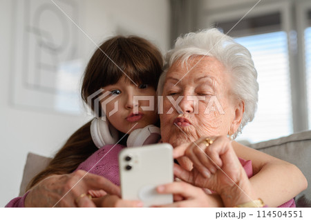 Grandmother with cute girl taking selfie with smartphone, making funny face. Portrait of an elderly woman spending time with granddaughter. 114504551