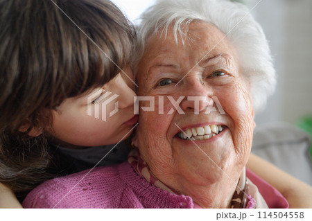 Cute girl hugging and kissing gradmother on cheek from behind. Portrait of an elderly woman spending time with granddaughter. 114504558