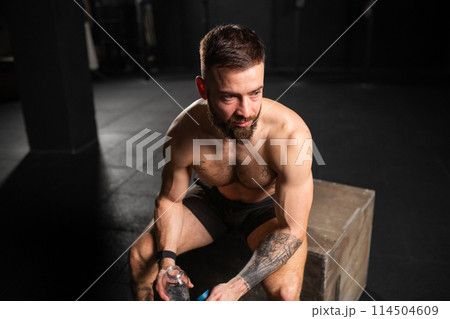 Man resting after exercise, drinking water from bottle, sitting, wearing short with bare chest. Routine workout for physical and mental health. 114504609