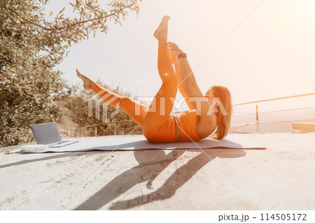 Fitness woman sea. Happy middle aged woman in orange sportswear exercises morning outdoors on yoga mat with laptop in park over ocean beach. Female fitness pilates yoga routine. Healthy lifestyle. Fitness woman sea. Happy middle aged woman in orange sportswear exercises morning outdoors on yoga mat with laptop in park over ocean beach. Female fitness pilates yoga routine. Healthy lifestyle. 114505172