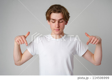 Young Man in White T-Shirt Pointing Downwards With Both Hands Against Plain Background 114506999
