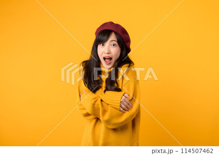 Surprise young Asian woman in her 30s, crossing her arms on her chest, wearing a yellow sweater and red beret against a yellow background. 114507462