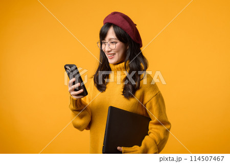 A young Asian woman, in a yellow sweater and red beret, confidently works on her laptop and smartphone against a vibrant yellow background. 114507467