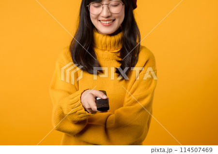 Close-up of a young Asian woman in her 30s, wearing a yellow sweater, happily changing channels on the TV with a remote control. 114507469