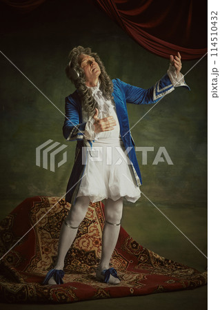 Overjoyed man dressed in richly detailed baroque costume listening music in headphones against vintage studio background. 114510432