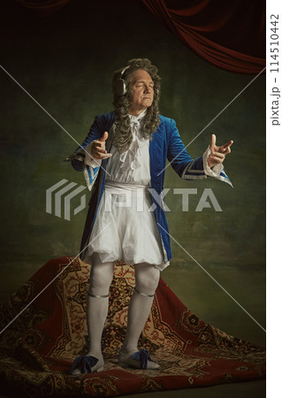 Elderly man dressed in richly detailed baroque costume with joyful expression listening music in headphones against vintage studio background. Elderly man dressed in richly detailed baroque costume with joyful expression listening music in headphones against vintage studio background. 114510442