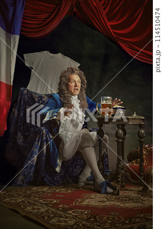 Elderly man, dressed as king in old-fashioned attire drink beer and watching sport channel on TV against vintage studio background. Elderly man, dressed as king in old-fashioned attire drink beer and watching sport channel on TV against vintage studio background. 114510474