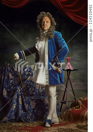 Full-length portrait of elderly man dressed as medieval person, aristocrat with confident expression against vintage studio background. Full-length portrait of elderly man dressed as medieval person, aristocrat with confident expression against vintage studio background. 114510492